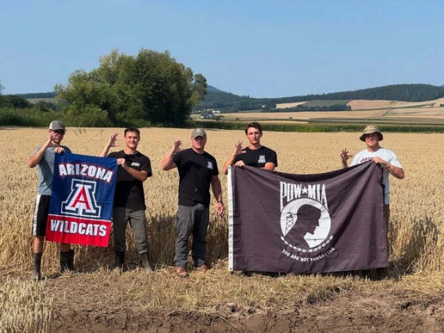Five people stand in a golden wheat field holding two flags—one red, white, and blue Arizona Wildcats flag and one black POW*MIA flag with the words “You Are Not Forgotten.” They wear casual outdoor clothing, some with boots and hats, and make the University of Arizona “Wildcat” hand sign against a backdrop of rolling farmland and blue sky.