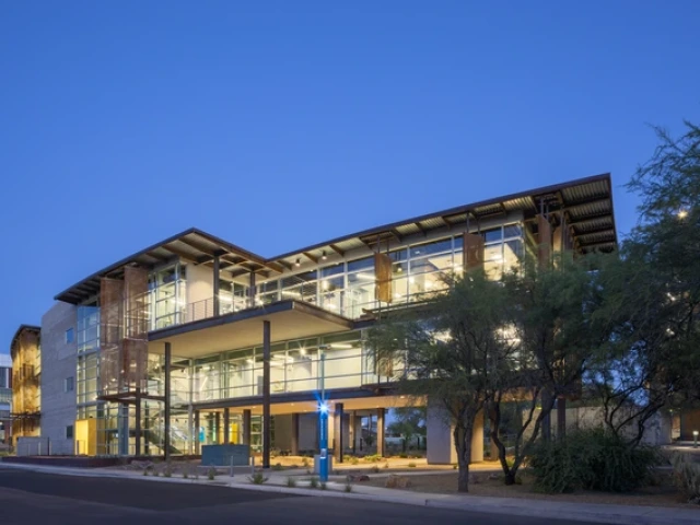 Modern three-story building with large glass windows, lit from inside at dusk, surrounded by trees and landscaping, with a blue emergency call box in front.
