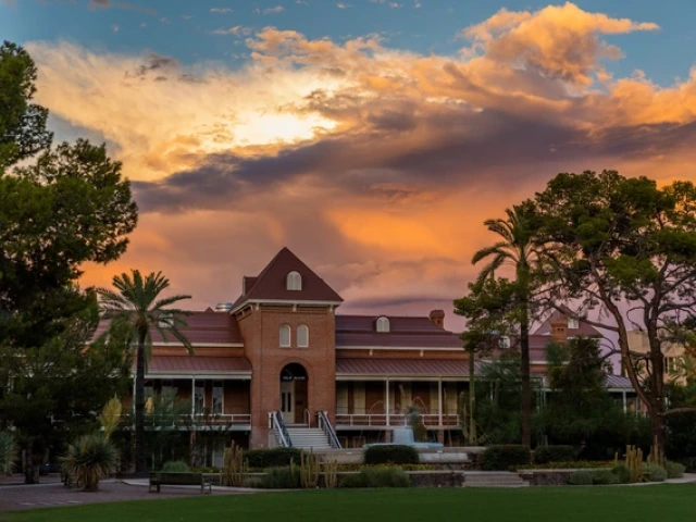 A photo of Old Main on the University of Arizona campus, a historic brick building with a prominent triangular roof and several chimneys, set against a vibrant sky. The sky is filled with dramatic clouds, transitioning from soft yellows and oranges to deep purples and blues, indicating either sunrise or sunset. In the foreground, well-maintained greenery includes palm trees, shrubs, and a neat lawn.