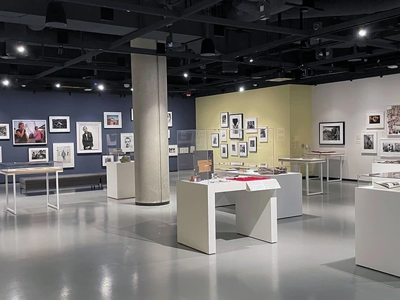 A modern gallery space at the Center for Creative Photography featuring an exhibition of framed photographs and archival materials. The walls are painted in dark blue, soft yellow, and white, with artworks arranged in salon-style groupings. Display cases throughout the room contain books, prints, and other photographic items under glass.