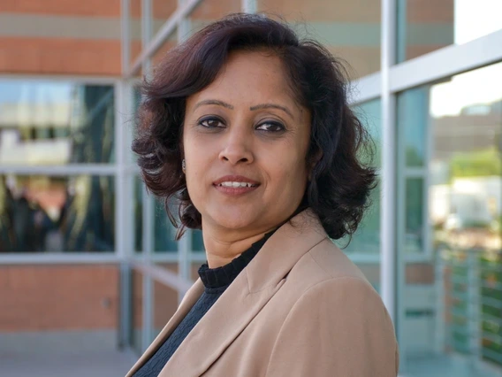 A confident woman with short, wavy dark hair stands in front of a modern glass and brick building. She wears a beige blazer over a black top and gazes calmly into the camera, with natural daylight highlighting her professional presence.