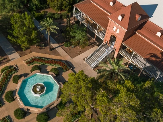 Aerial view of a historic red-brick building with a brown metal roof and a prominent staircase leading to the entrance. In front of the building, an octagonal water fountain with a central pedestal is surrounded by paved walkways, manicured gardens, and green trees. The scene is bathed in warm sunlight, casting long shadows across the landscape.