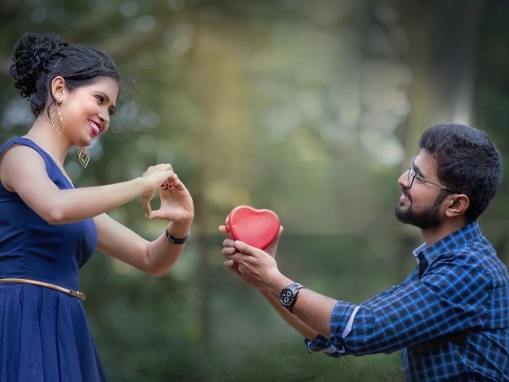 A man on bended knee offers a heart-shaped gift to a woman. 