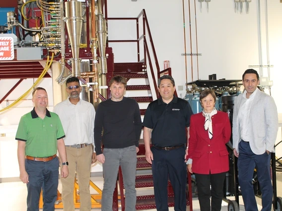 A group of six professionals stands in front of an industrial research setup with metal pipes, wiring, and machinery. The team members wear a mix of business casual and professional attire, with some dressed in polo shirts, blazers, and button-down shirts. A sign in the background reads 'ABSOLUTELY NO STORAGE BELOW,' emphasizing the safety precautions in the high-tech facility.