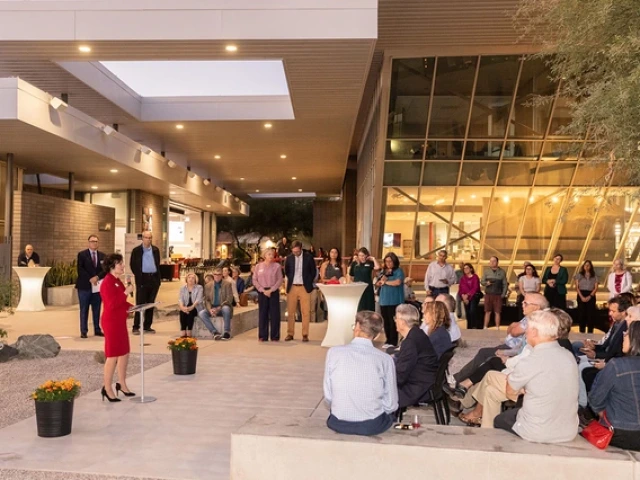 A woman speaks at a podium to an audience during an outdoor evening event at a modern university building.