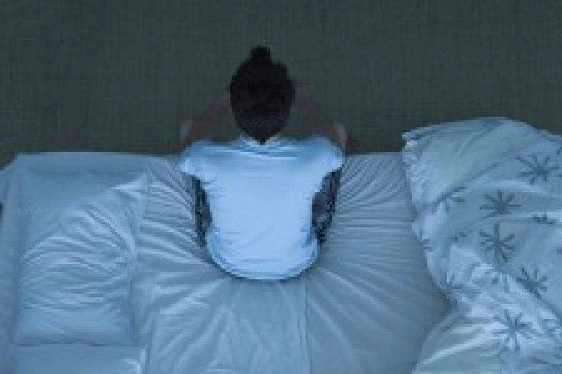 A man in a white shirt and grey sweat pants sits at the edge of a bed. 