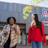 Two students walk and chat outside the University of Arizona Global Center. One student, wearing a plaid shirt and glasses, gestures while holding a phone, while the other, in a red sweatshirt and backpack, smiles. The modern building with the university's "A" logo is visible in the background.