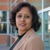 A confident woman with short, wavy dark hair stands in front of a modern glass and brick building. She wears a beige blazer over a black top and gazes calmly into the camera, with natural daylight highlighting her professional presence.