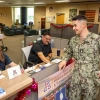 A U.S. Navy service member in a camouflage uniform chats with two men seated at a front desk in a military student support center. The desk is decorated with American flags, brochures, and a sign reading "Veterans Education & Transition Services" from the University of Arizona. The setting is an inviting space with couches, decorations, and a whiteboard that says, "Please sign in here."